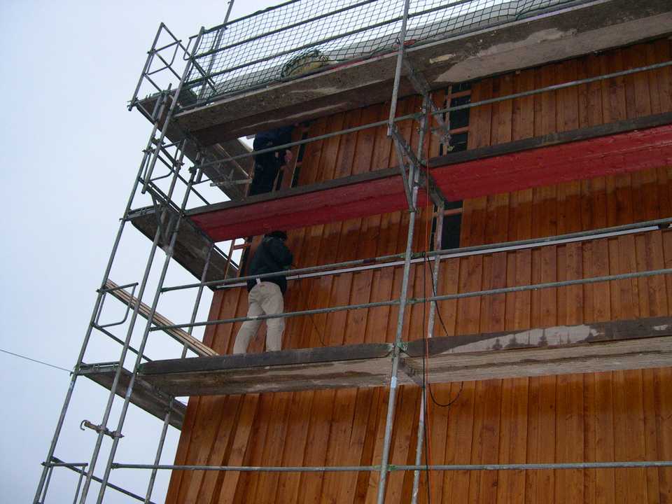 An vier Flächen der Hallenfassade wird eine großflächige Holzverschalung an die Wand montiert. Direkt an die zur südlichen Giebelwand anliegenden Fläche sind die Arbeiten bereits weit fortgeschritten.