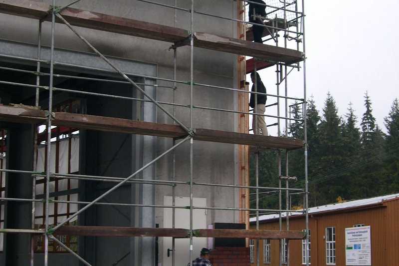 An der Giebelseite der Halle mit den Gleiseinfahrten wird die die Klinkerwand aufgebaut, an der Seitenwand arbeiten die Zimmerleute beim Aufbau der Holzverblendung.